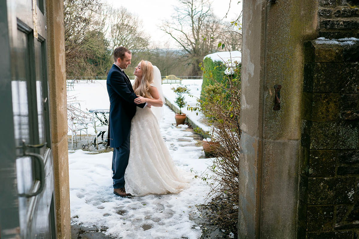 A newly married couple in traditional dress stand in the snow, looking at each other at Hassop Hall in Derbyshire.